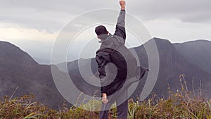 Young male hiker in raincoat with backpack reaching up top of mountain and raised hands. Man tourist standing on the