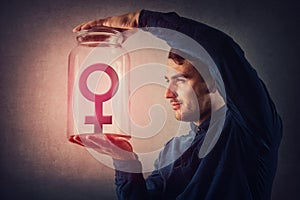 Young male, malefic look holding a glass jar with female gender symbol inside as captive. Man pretending to be superior to woman, photo
