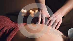 Young male massage therapist does back massage to a woman with a tattoo in a massage room with dim lights on the