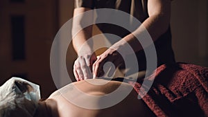 Young male massage therapist does back massage to a woman with a tattoo in a massage room with dim lights on the