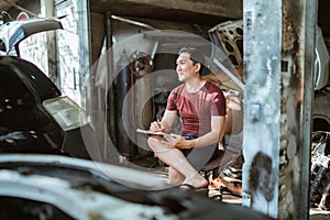 young male mechanic sits recalling while writing a list of car repairs
