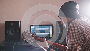 Young male musician composes and records soundtrack playing the guitar using computer, headphones and keyboard