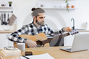 Young male musician tuning guitar using laptop on desk in modern apartment.