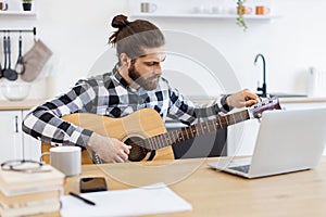 Young male musician tuning guitar using laptop on desk in modern apartment.