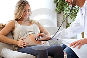 Young male obstetrician doctor with stethoscope listening to beautiful pregnant woman baby heartbeat.