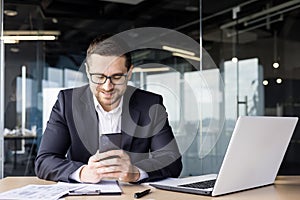 A young male office worker sits at a desk and uses the phone, smilingly typing messages, reading news, playing online