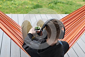 Young male person enjoys listening music in headset from cell phone and resting in hammock outdoors.