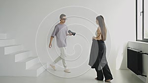 Young male photographer with glasses shows model girl poses for photo on stairs in bright studio with large windows