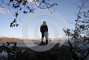 A young male photographer silhouette over a huge rock capturing a lake landscape