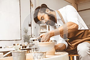 A young male potter is engaged in craft in his workshop on a potter`s wheel and makes a clay product