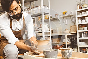 A young male potter is engaged in craft in his workshop on a potter`s wheel and makes a clay product