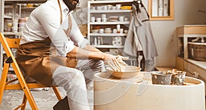 A young male potter is engaged in craft in his workshop on a potter`s wheel and makes a clay product