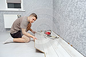 Young male repairman is laying panel of laminate floor indoors on the knees