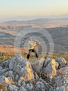 Young male saxophonist in black pants and yellow jacket, stands at the top of mountain against background of valley and holds