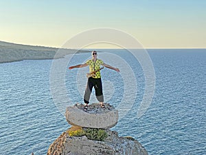 A young male saxophonist in black wide pants and a yellow T-shirt, stands at the top of a cliff against the background of the sea