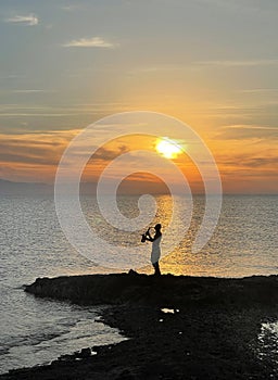 Young male saxophonist stands against background of sea and sunset, holds saxophone in his hands. Beautiful sunset on sea, sky.