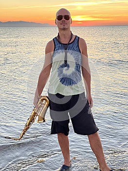 Young male saxophonist stands with his feet in sea water, holds saxophone in his hands. Beautiful sunset on sea, sky. Musician,