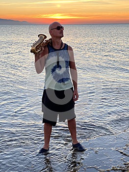 Young male saxophonist stands with his feet in sea water, holds saxophone in his hands. Beautiful sunset on sea, sky. Musician,