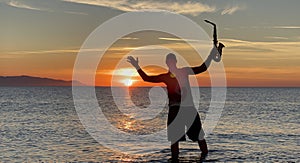 Young male saxophonist stands with his feet in sea water, holds saxophone in his hands, looks at sunset. Beautiful sunset on sea,