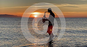 Young male saxophonist stands with his feet in sea water, holds saxophone in his hands, looks at sunset. Beautiful sunset on sea,