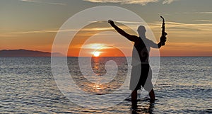 Young male saxophonist stands with his feet in sea water, holds saxophone in his hands, looks at sunset. Beautiful sunset on sea,