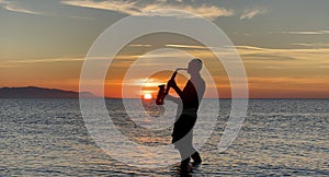 Young male saxophonist stands with his feet in sea water, holds saxophone in his hands, looks at sunset. Beautiful sunset on sea,