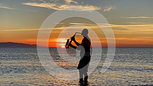Young male saxophonist stands with his feet in sea water, holds saxophone in his hands, looks at sunset. Beautiful sunset on sea,