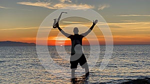 Young male saxophonist stands with his feet in sea water, holds saxophone in his hands, looks at sunset. Beautiful sunset on sea,