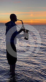 Young male saxophonist stands with his feet in sea water, holds saxophone in his hands, looks at sunset. Beautiful sunset on sea,