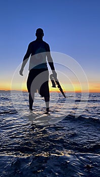 Young male saxophonist stands with his feet in sea water, holds saxophone in his hands, looks at sunset. Beautiful sunset on sea,