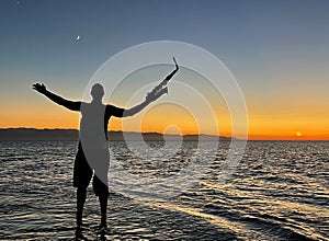 Young male saxophonist stands with his feet in sea water, holds saxophone in his hands, looks at sunset. Beautiful sunset on sea,