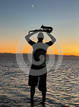 Young male saxophonist stands with his feet in sea water, holds saxophone in his hands, looks at sunset. Beautiful sunset on sea,