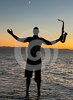 Young male saxophonist stands with his feet in sea water, holds saxophone in his hands, looks at sunset. Beautiful sunset on sea,