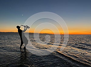 Young male saxophonist stands with his feet in sea water, holds saxophone in his hands, looks at sunset. Beautiful sunset on sea,