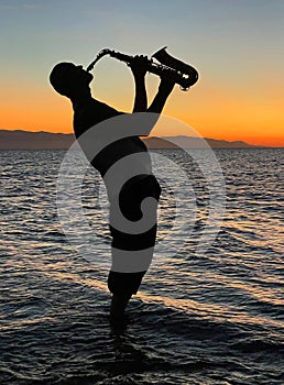 Young male saxophonist stands with his feet in sea water, holds saxophone in his hands, looks at sunset. Beautiful sunset on sea,