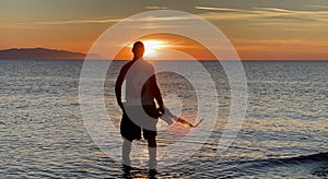 Young male saxophonist stands with his feet in sea water, holds saxophone in his hands, looks at sunset. Beautiful sunset on sea,