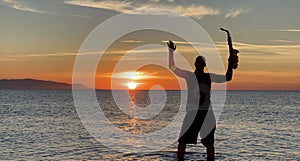 Young male saxophonist stands with his feet in sea water, holds saxophone in his hands, looks at sunset. Beautiful sunset on sea,