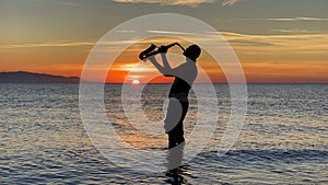 Young male saxophonist stands with his feet in sea water, holds saxophone in his hands, looks at sunset. Beautiful sunset on sea,