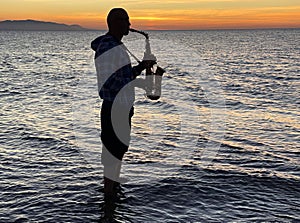 Young male saxophonist stands with his feet in sea water, holds saxophone in his hands, looks at sunset. Beautiful sunset on sea,