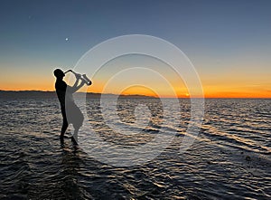 Young male saxophonist stands with his feet in sea water, holds saxophone in his hands, looks at sunset. Beautiful sunset on sea,