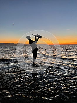 Young male saxophonist stands with his feet in sea water, holds saxophone in his hands, looks at sunset. Beautiful sunset on sea,