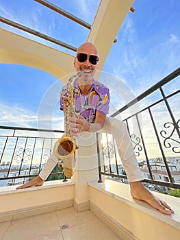 A young male saxophonist in white pants and a purple T-shirt stands on the balcony of a hotel in Cyprus and holds a saxophone