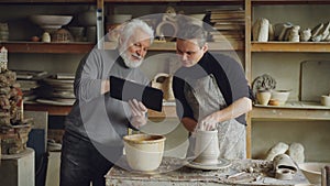 Young male sculptor is making clay figure on throwing wheel while his father is showing him tablet, men are discussing