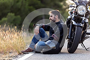 Young male sitting on the road, leaning on his motorcycle while waiting to the roadside assistance to repair the motorbike