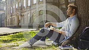 Young male sitting under tree with book looking around, having pleasant thoughts
