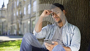 Young male sitting under tree, reading information on cellphone, upsetting news