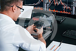 Young male stock brocker works indoors in the office. Many of displays