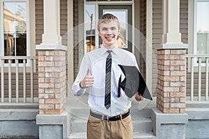 Young male student holding a diploma and a graduation cap in front of a house.