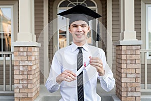 Young male student holding a diploma while wearing a graduation cap in front of a house.