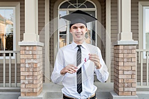 Young male student holding a diploma while wearing a graduation cap in front of a house.